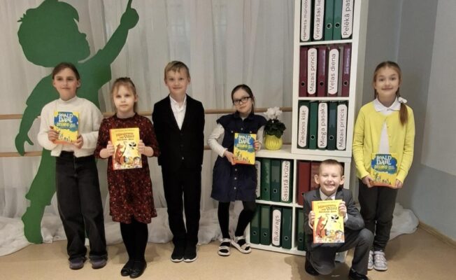 Seši smaidīgi bērni, pieci stāv un viens tup, tur rokās grāmatas. Vairākām grāmatām uz vāka redzams uzraksts Roald Dahl. Aiz viņiem atrodas balts grāmatu plaukts ar mapēm.