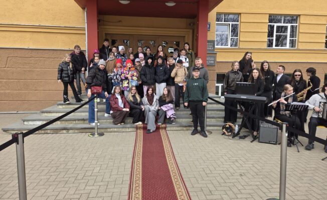 Liela jauniešu grupa stāv un sēž uz dzeltenas ēkas kāpnēm un pie sarkana paklāja. Labajā pusē mūzikas ansamblis spēlē flautu, saksofonu, akordeonu un sintezatoru. Uz ēkas plāksnes redzams uzraksts MŪZIKAS SKOLA.