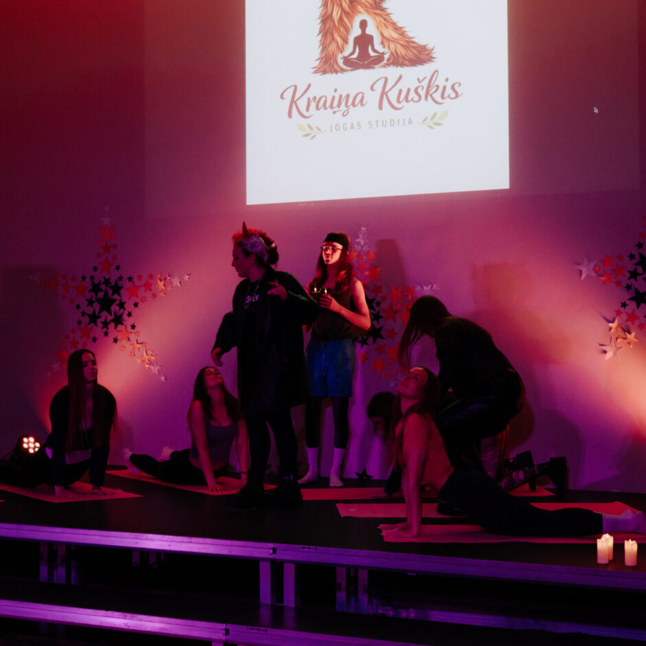 Uz skatuves ar purpursarkanu apgaismojumu vairāki cilvēki veic jogas pozas uz sarkaniem paklājiņiem, kamēr daži stāv.