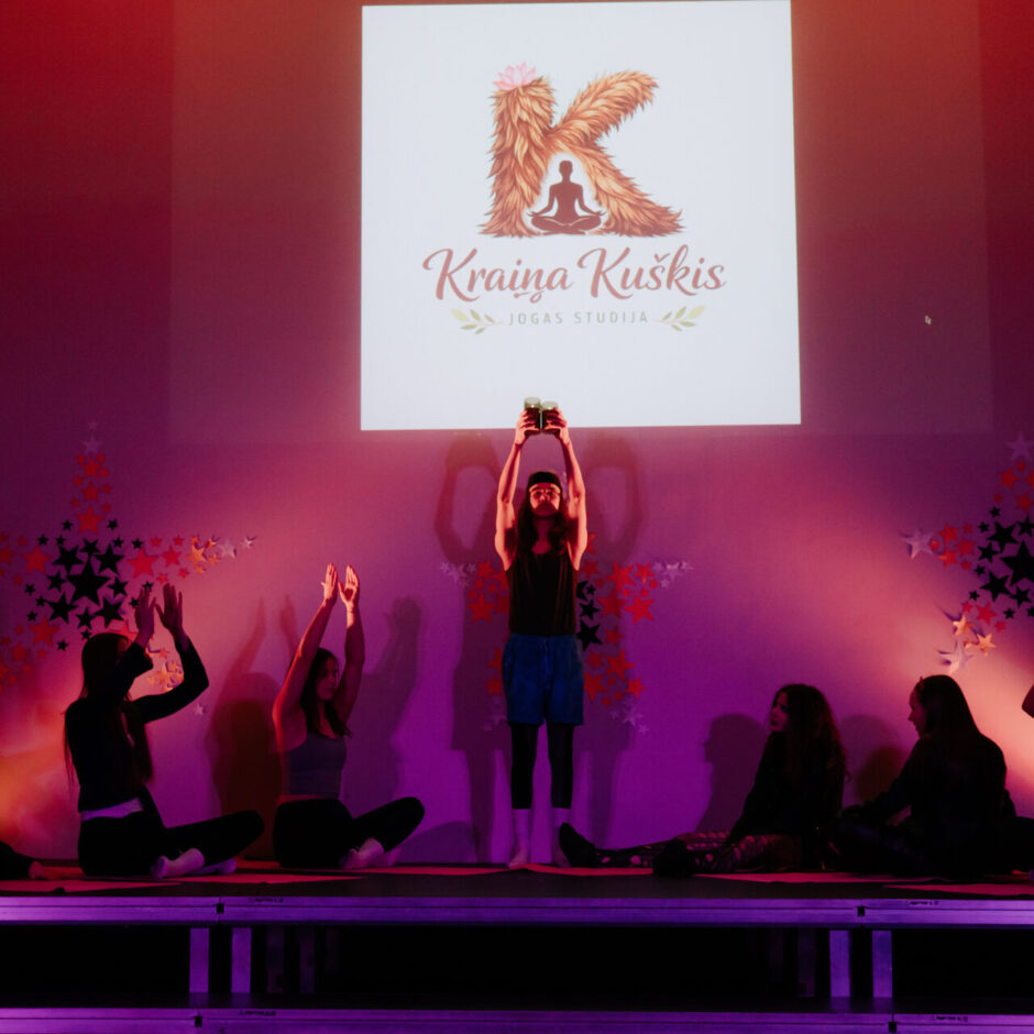 Cilvēku grupa nodarbojas ar jogu blāvi apgaismotā telpā ar purpursarkanām un sarkanām gaismām.