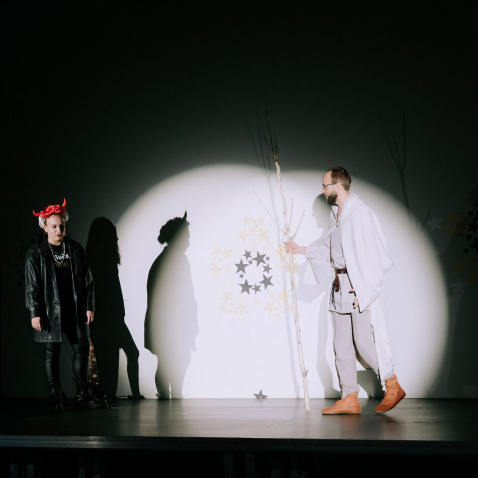 Teātra uzvedumā uz tumšas skatuves prožektors izgaismo vīrieti baltā halātā ar zaru rokā. Pa kreisi tumsā stāv divas personas, no kurām viena ir ar sarkaniem ragiem un melnā ādas jakā. Fonā redzamas zvaigznes un koku zaru silueti.