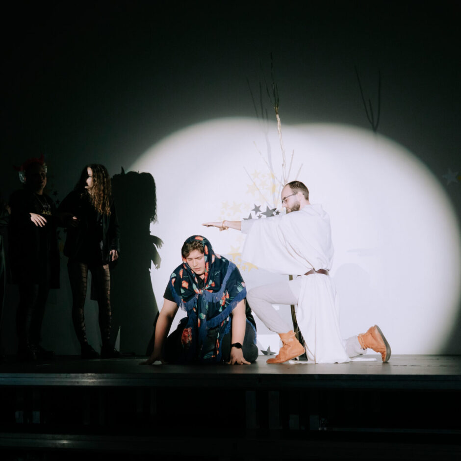 Uz skatuves notiek izrāde. Trīs jaunieši tumšās drēbēs stāv pa kreisi ēnā,