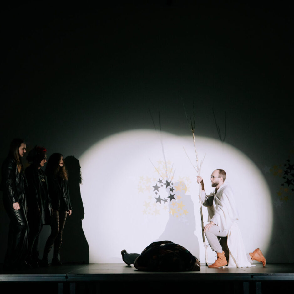 Uz skatuves, ko izgaismo prožektors, pa labi nometies ceļos vīrietis gaišā apģērbā, turot koka zaru.