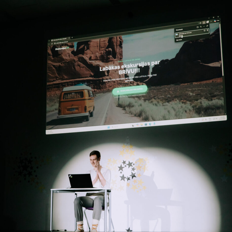 Jauns vīrietis sēž uz skatuves pie galda ar atvērtu klēpjdatoru, smaidot un skatoties uz ekrānu. Aiz viņa uz sienas tiek projicēta tīmekļa vietne