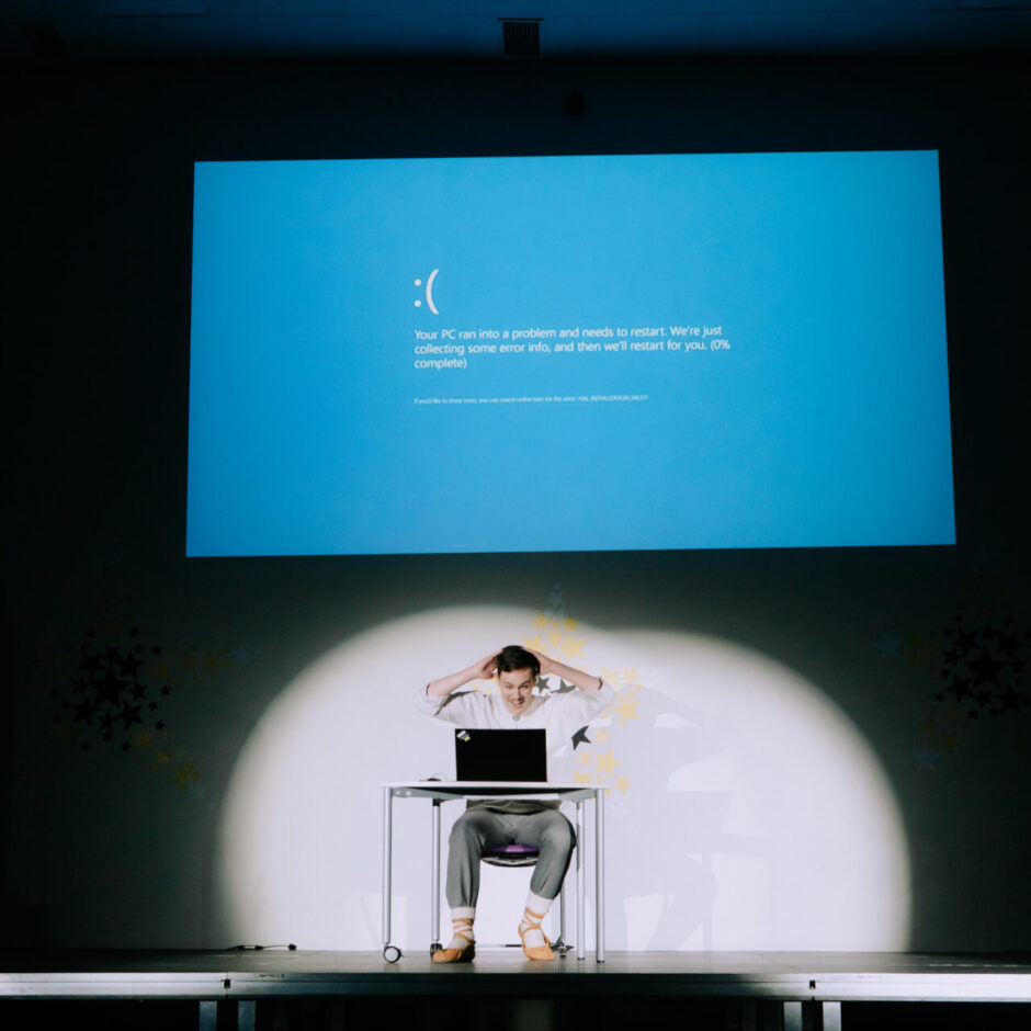 Vīrietis sēž pie galda uz skatuves spožā gaismā, izmisis satvēris galvu, skatoties uz klēpjdatoru. Aiz viņa uz liela ekrāna tiek projicēts zils kļūdas ekrāns ar tekstu: 
