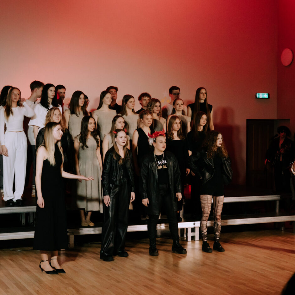 Liela pusaudžu un jauniešu grupa uz skatuves sarkanīgā apgaismojumā. Fonā daudzi stāv rindās uz paaugstinājumiem. Priekšplānā pa kreisi meitene melnā kleitā žestikulē, centrā trīs meitenes melnos tērpos.