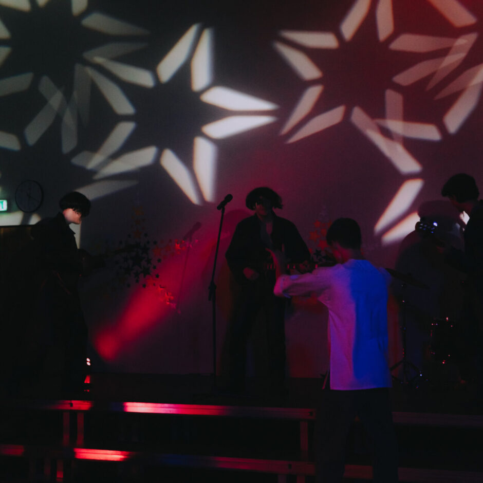 Mūzikas grupa uz skatuves tumšā telpā ar sarkanu un zilu skatuves apgaismojumu un zvaigžņu veida projekcijām uz sienas.