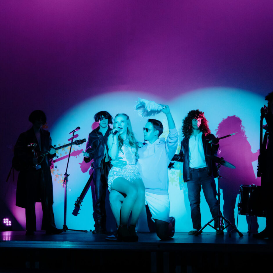 Skatuves priekšplānā blondīne mirdzošā kleitā dzied mikrofonā, kamēr uz ceļa nometies vīrietis saulesbrillēs virs viņas galvas tur baltu putekļu slotiņu. Fonā redzami vairāki mūziķi ar ģitārām un bungām, izgaismoti violetos un tirkīzzilos toņos.