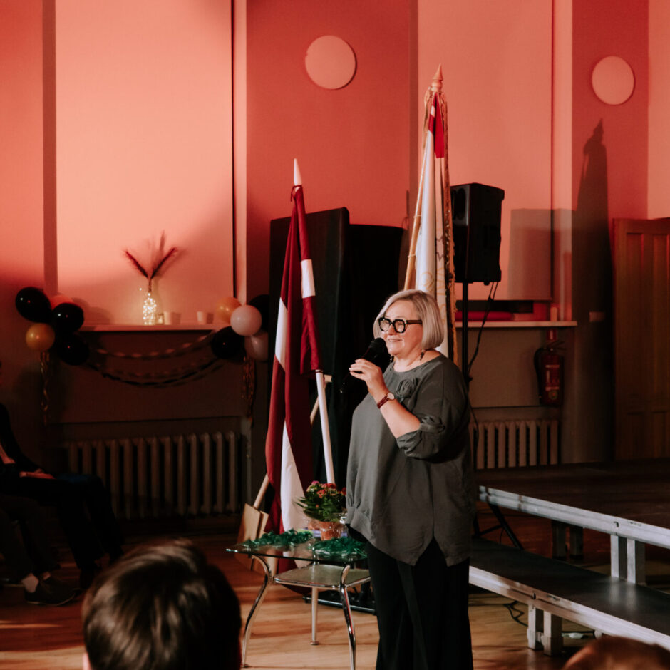 Sieviete ar brillēm runā mikrofonā, stāvot uz koka grīdas izgaismotā telpā. Aiz viņas atrodas Latvijas karogs un cits ceremonijas karogs, bet priekšplānā redzamas auditorijas muguras.