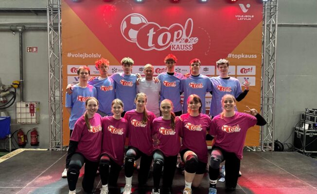 Divpadsmit volejbolistu grupa – piecas meitenes ceļos priekšplānā rozā krekliņos un seši zēni aizmugurē zilos krekliņos – pozē fotogrāfijai kopā ar pieaugušu treneri baltā kreklā. Viņi stāv sarkana un oranža banera priekšā .