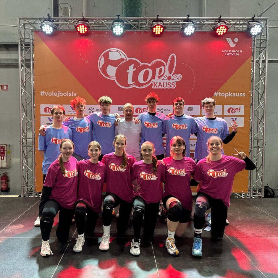 Divpadsmit volejbolistu grupa – piecas meitenes ceļos priekšplānā rozā krekliņos un seši zēni aizmugurē zilos krekliņos – pozē fotogrāfijai kopā ar pieaugušu treneri baltā kreklā. Viņi stāv sarkana un oranža banera priekšā .