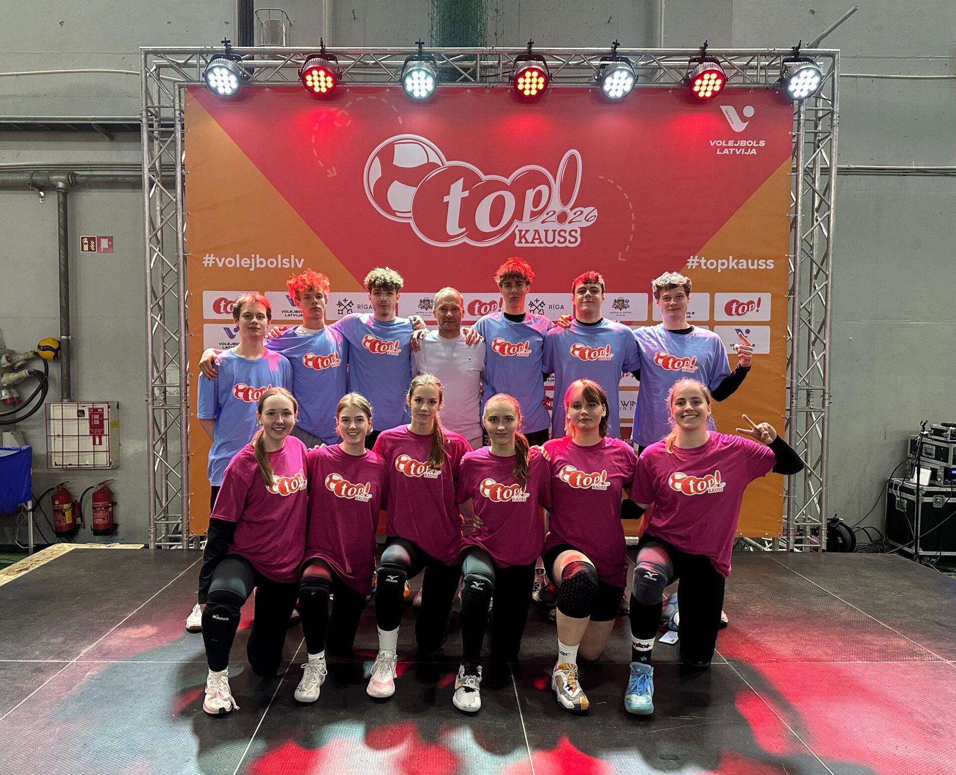 Divpadsmit volejbolistu grupa – piecas meitenes ceļos priekšplānā rozā krekliņos un seši zēni aizmugurē zilos krekliņos – pozē fotogrāfijai kopā ar pieaugušu treneri baltā kreklā. Viņi stāv sarkana un oranža banera priekšā .